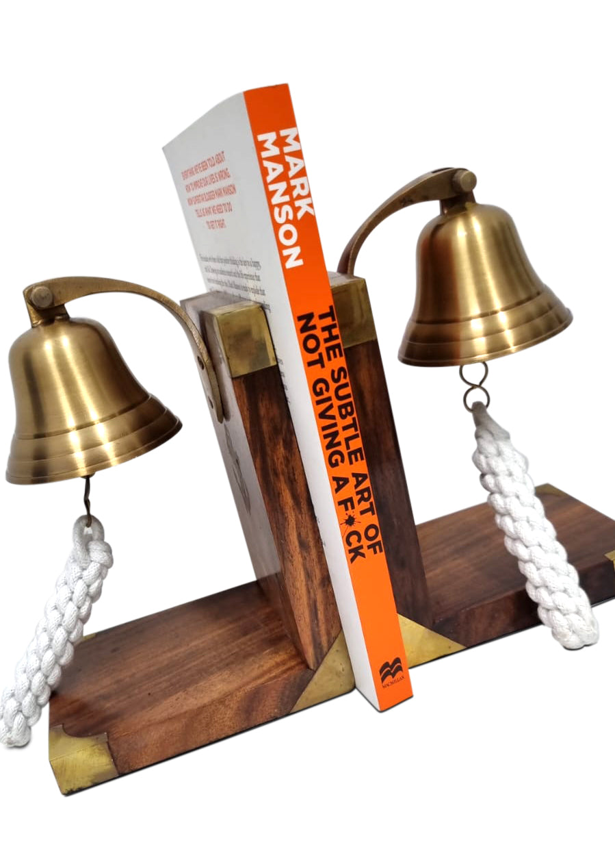 Two book ends of Brass marine bell on a L shaped wood base with a book between them Title is The subtle art of not giving a F*ck by Mark Manson.