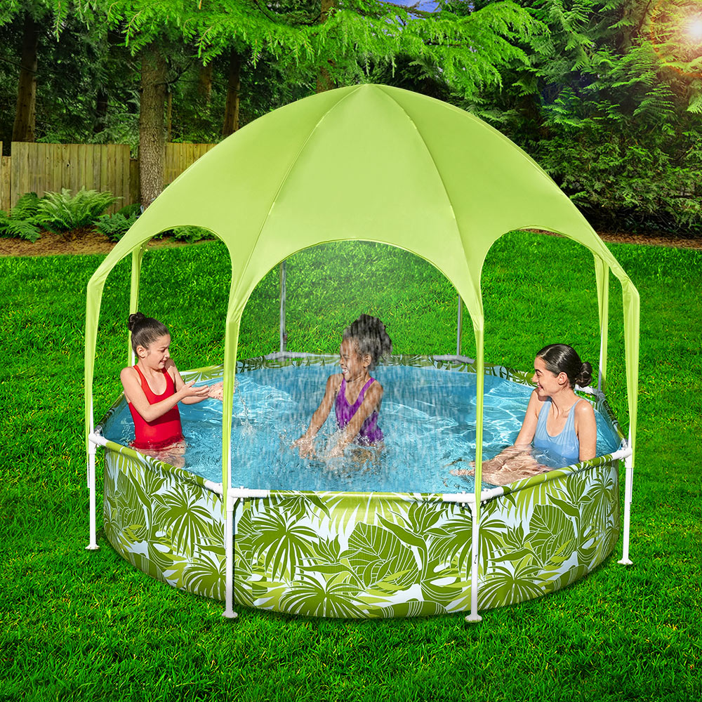 A lady and 2 little children splashing in an above ground swimming pool that is round with a fixed sun shade and has a green palm leaf image design on the nice green grass of home. 