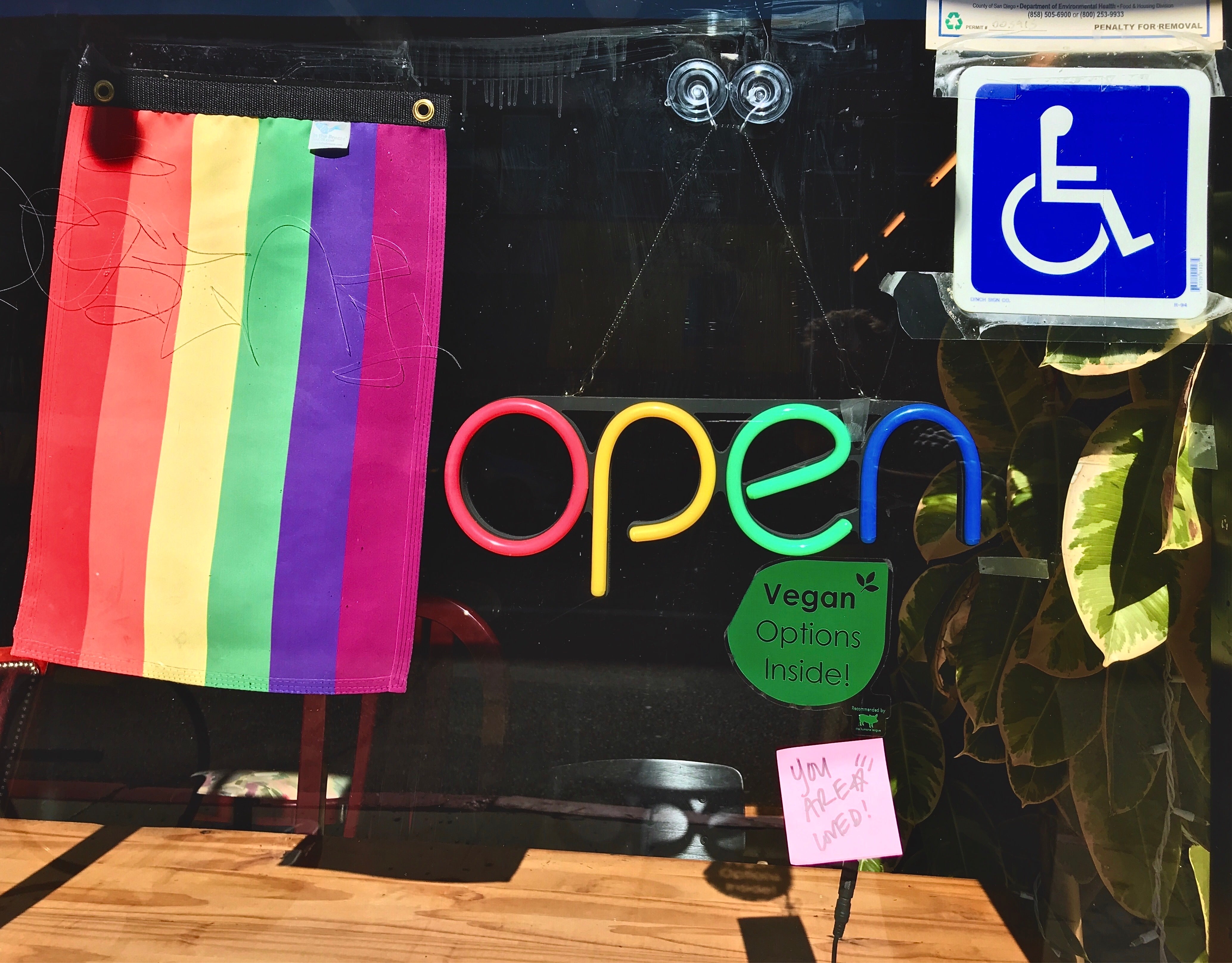 Rainbow flag and open sign with a disabled wheelchair sticker with vegan options you are loved at a cafe window and table.